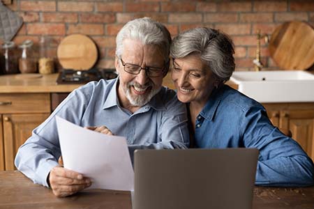  Lächelndes Rentnerpaar sitzt vor einem Laptop und hält ein Schreiben in Händen